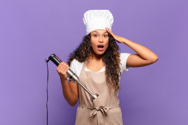 Young hispanic woman looking happy, astonished and surprised, smiling and realizing amazing and incredible good news. chef concept