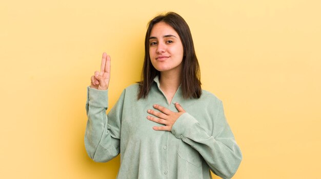 Young hispanic woman looking happy confident and trustworthy smiling and showing victory sign with a positive attitude