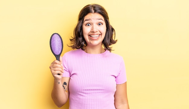 Young hispanic woman looking happy and pleasantly surprised. hairbrush concept