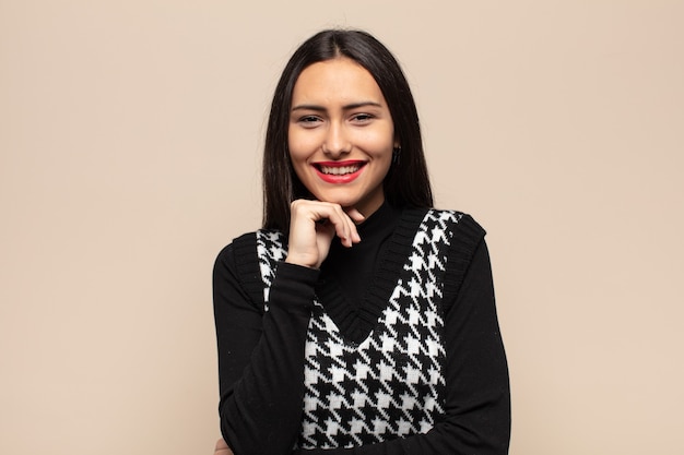 Young hispanic woman looking happy and smiling with hand on chin, wondering or asking a question, comparing options