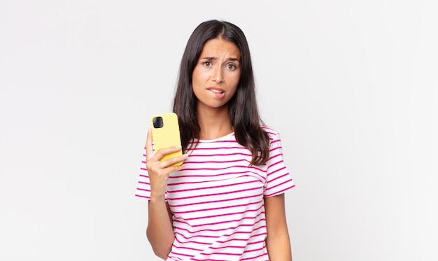 Young hispanic woman looking puzzled and confused and holding a smartphone