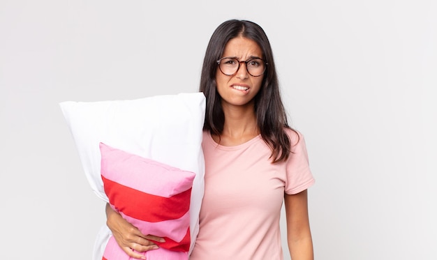 Young hispanic woman looking puzzled and confused wearing pajamas and holding a pillow