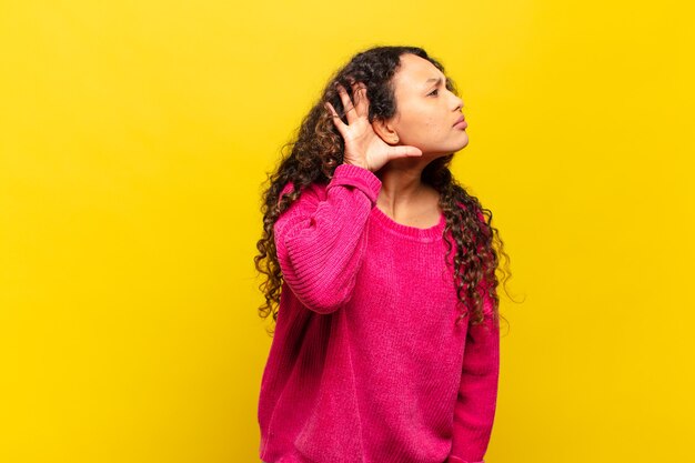 Young hispanic woman looking serious and curious, listening, trying to hear a secret conversation or gossip, eavesdropping