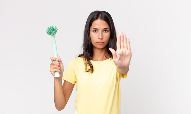 Young hispanic woman looking serious showing open palm making stop gesture and holding a dish cleaning brush