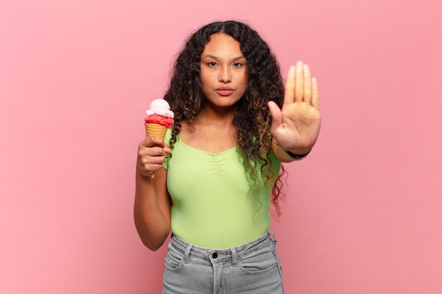 Young hispanic woman looking serious, stern, displeased and angry showing open palm making stop gesture. ice cream concept