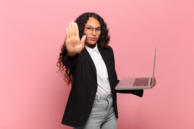 真面目で、厳しく、不機嫌で、怒っている若いヒスパニック系の女性が、手のひらを開いてジェスチャーを止めているのを見せています。ノートパソコンのコンセプト