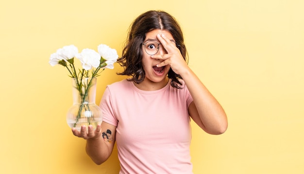 Young hispanic woman looking shocked, scared or terrified, covering face with hand. flowers pot concept