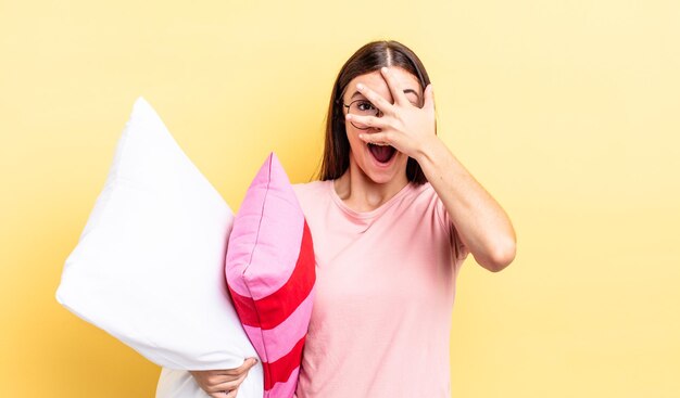 Young hispanic woman looking shocked, scared or terrified, covering face with hand. pajamas and pillow concept