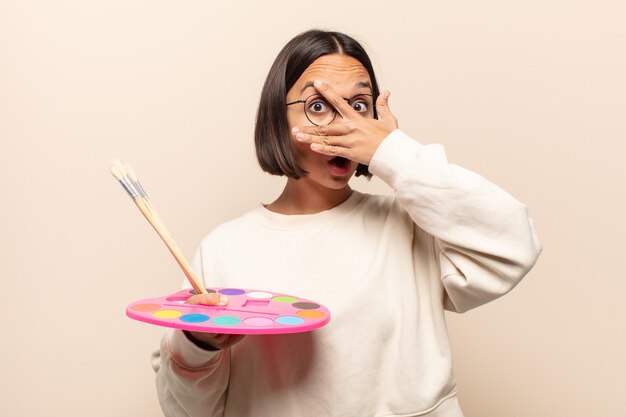 Young hispanic woman looking shocked, scared or terrified, covering face with hand and peeking between fingers