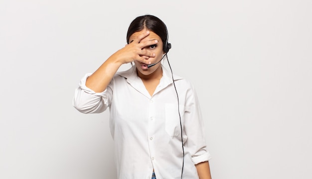 Young hispanic woman looking shocked, scared or terrified, covering face with hand and peeking between fingers