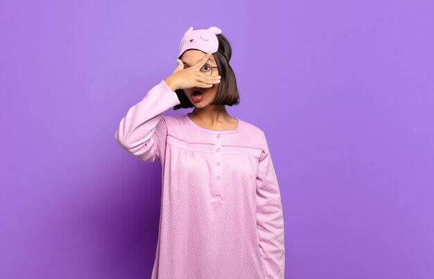 Young hispanic woman looking shocked, scared or terrified, covering face with hand and peeking between fingers