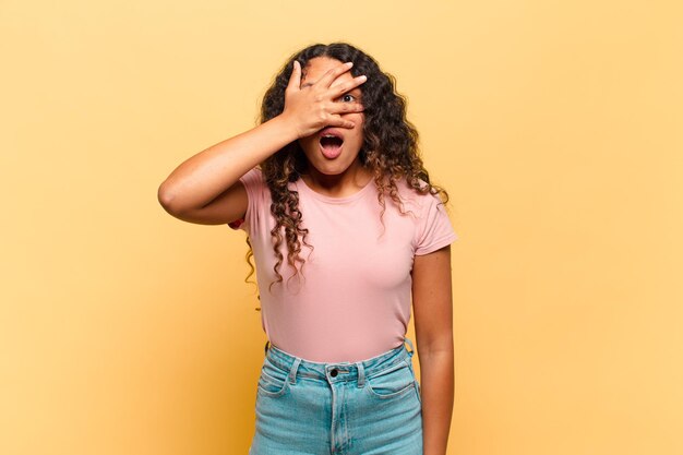 Young hispanic woman looking shocked, scared or terrified, covering face with hand and peeking between fingers