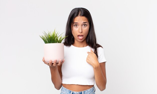 Young hispanic woman looking shocked and surprised with mouth wide open, pointing to self and holding a decorative house plant