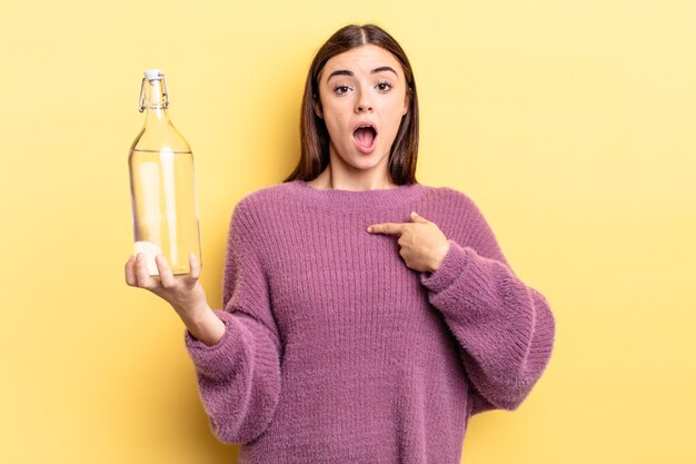 Young hispanic woman looking shocked and surprised with mouth wide open, pointing to self. water bottle concept