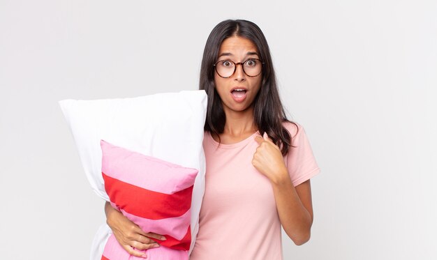 Young hispanic woman looking shocked and surprised with mouth wide open, pointing to self wearing pajamas and holding a pillow