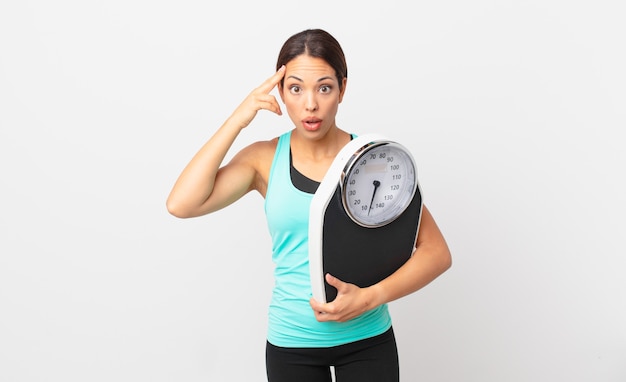 Young hispanic woman looking surprised, realizing a new thought, idea or concept and holding a scale