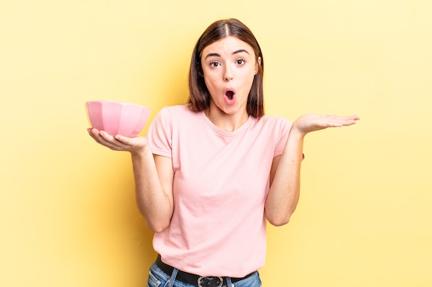 young hispanic woman looking surprised and shocked, with jaw dropped holding an object. empty bowl concept