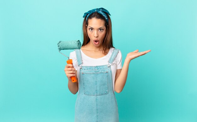 young hispanic woman looking surprised and shocked, with jaw dropped holding an object and painting a wall