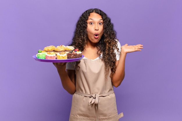 Young hispanic woman looking surprised and shocked, with jaw dropped holding an object with an open hand on the side. cooking cakes concept