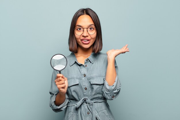 Young hispanic woman looking surprised and shocked, with jaw dropped holding an object with an open hand on the side