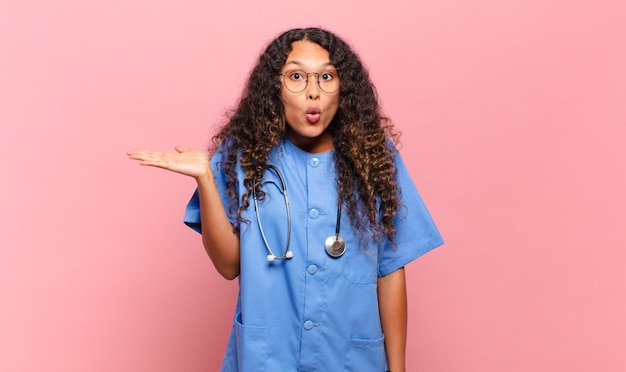 Young hispanic woman looking surprised and shocked, with jaw dropped holding an object with an open hand on the side.