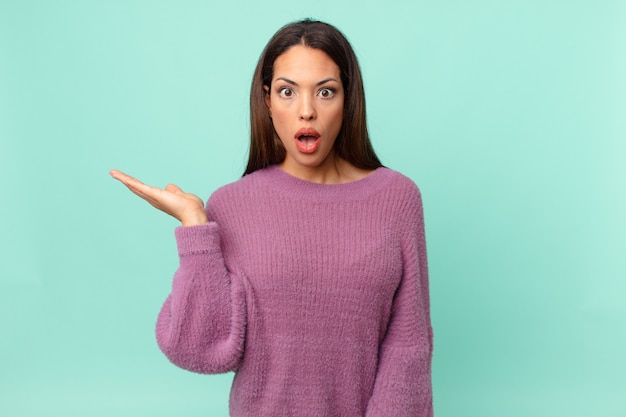 Young hispanic woman looking surprised and shocked, with jaw dropped holding an object