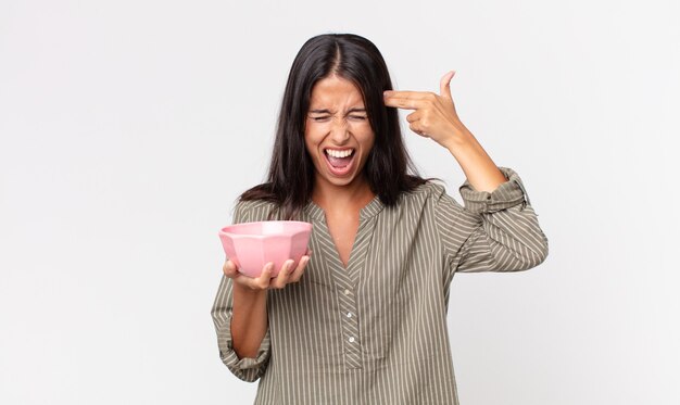 Young hispanic woman looking unhappy and stressed, suicide gesture making gun sign and holding an empty bowl or pot