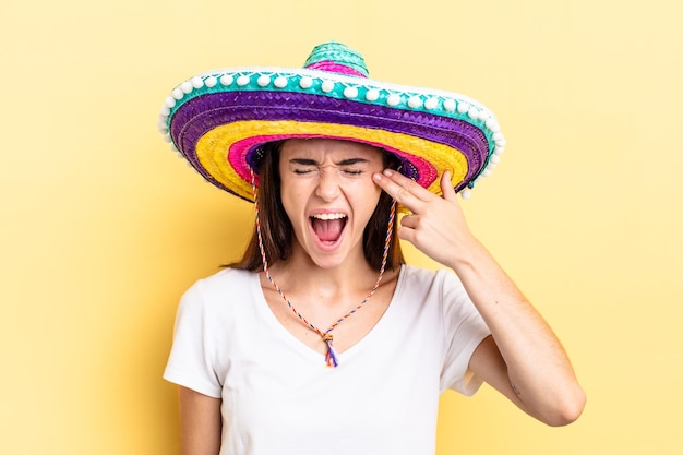 Молодая латиноамериканская женщина, выглядящая несчастной и подчеркнутой, жест самоубийства, делая знак пистолета. концепция мексиканской шляпы