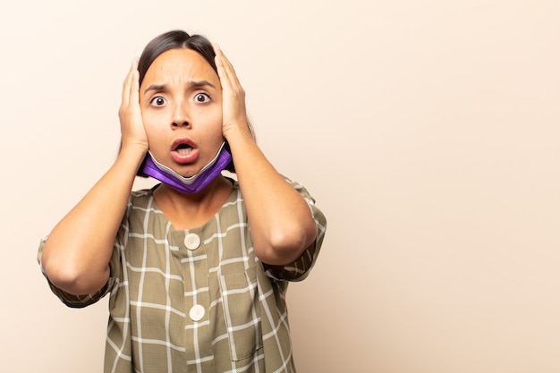 Young hispanic woman looking unpleasantly shocked, scared or worried, mouth wide open and covering both ears with hands