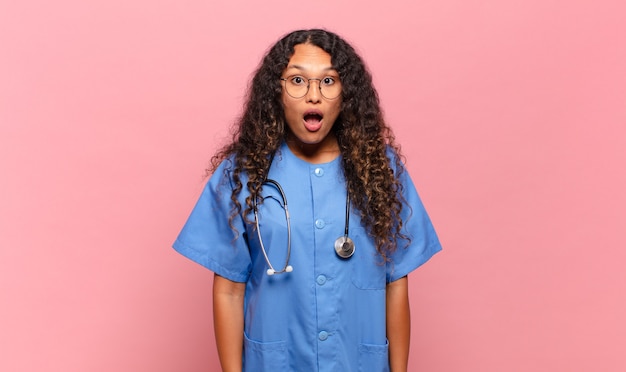 Young hispanic woman looking very shocked or surprised, staring with open mouth saying wow. nurse concept