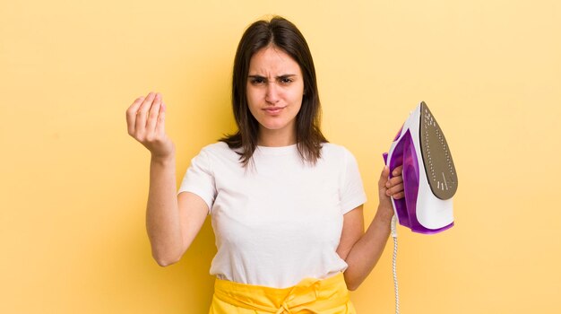 Young hispanic woman making capice or money gesture telling you to pay clothes iron concept