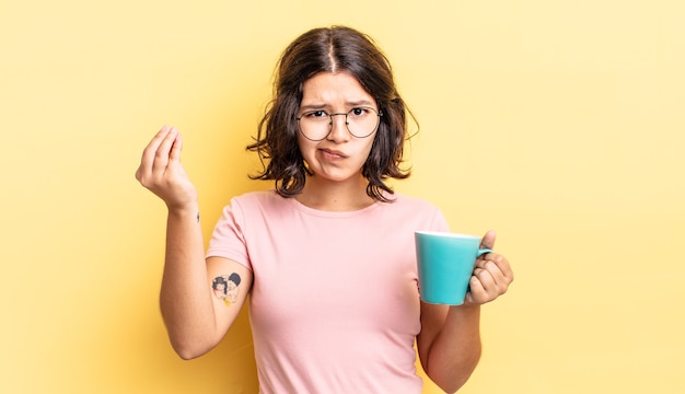 young hispanic woman making capice or money gesture, telling you to pay. coffee mug concept