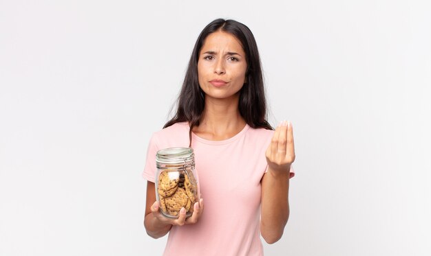 Young hispanic woman making capice or money gesture, telling you to pay and holding a cookies glass bottle