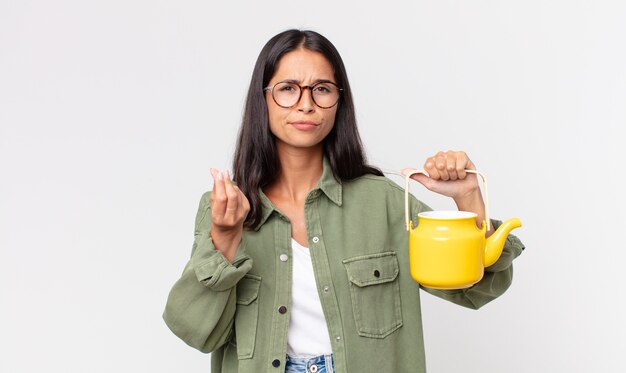 Young hispanic woman making capice or money gesture, telling you to pay and holding a tea pot