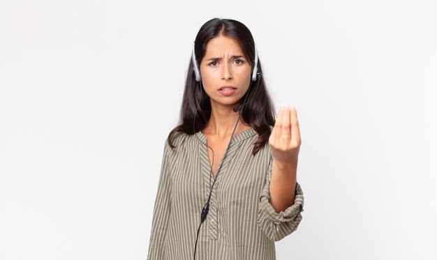 Young hispanic woman making capice or money gesture, telling you to pay with a headset. telemarketing concept