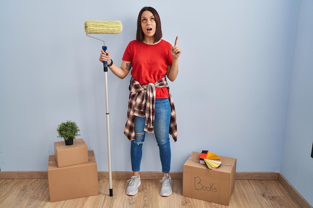 Young hispanic woman painting home walls with paint roller amazed and surprised looking up and pointing with fingers and raised arms.