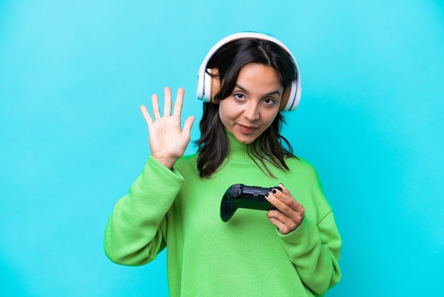 Giovane donna ispanica che gioca con un controller per videogiochi isolato su sfondo blu contando cinque con le dita