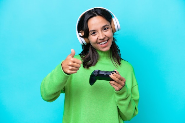 Young hispanic woman playing with a video game controller isolated on blue background with thumbs up because something good has happened