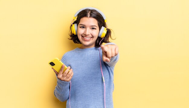 Giovane donna ispanica che punta alla telecamera scegliendo te. cuffie e concetto di smartphone