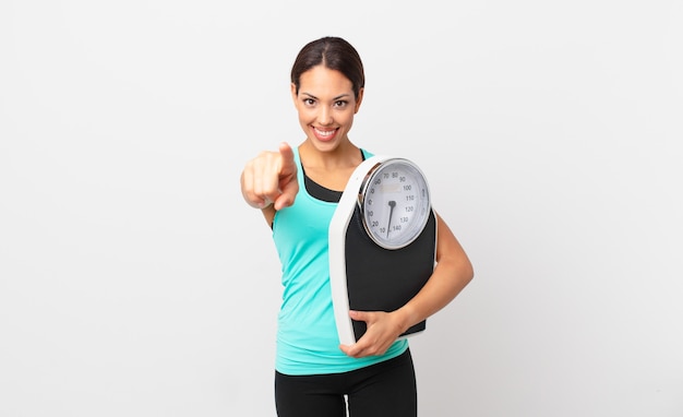 Young hispanic woman pointing at camera choosing you and holding a scale