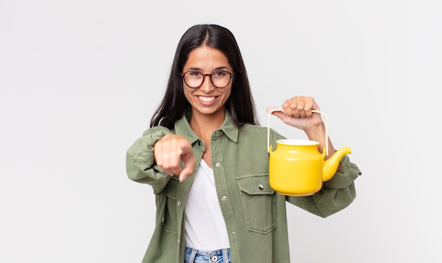 Young hispanic woman pointing at camera choosing you and holding a tea pot