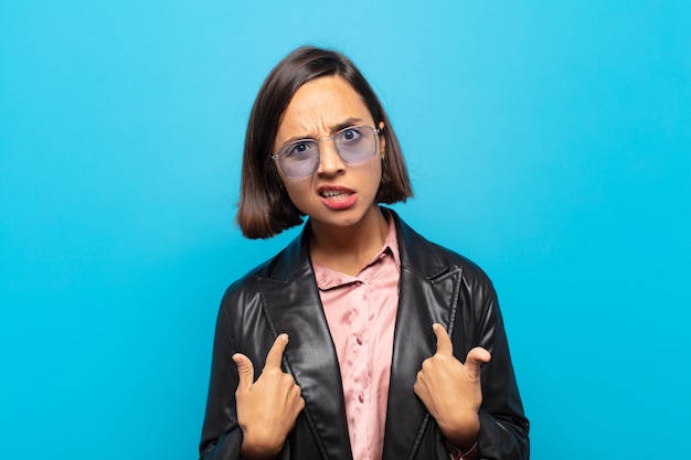 Young hispanic woman pointing to self with a confused and quizzical look, shocked and surprised to be chosen