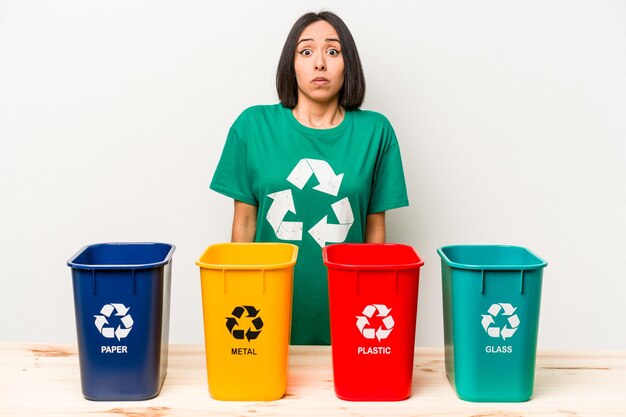 Young hispanic woman recycling isolated on white background shrugs shoulders and open eyes confused