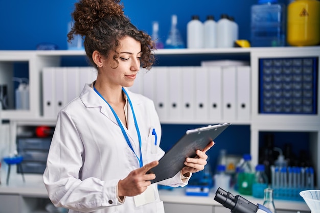 Young hispanic woman scientist smiling confident reading document at laboratory