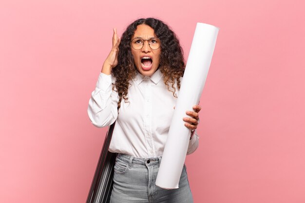 Young hispanic woman screaming with hands up in the air, feeling furious, frustrated, stressed and upset. architect concept