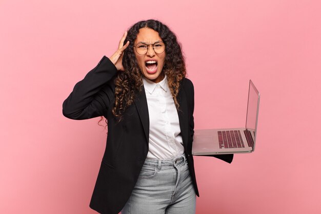 Young hispanic woman screaming with hands up in the air, feeling furious, frustrated, stressed and upset. laptop concept