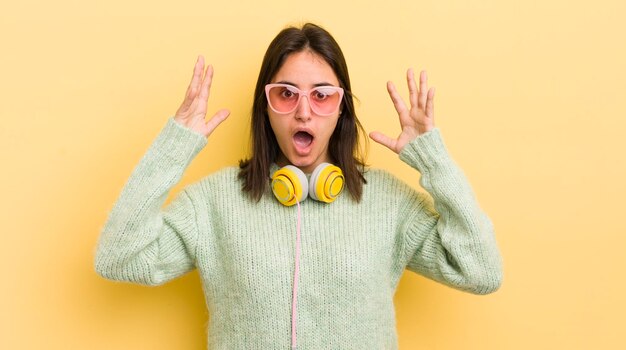 Photo young hispanic woman screaming with hands up in the air headphones and sunglasses concept