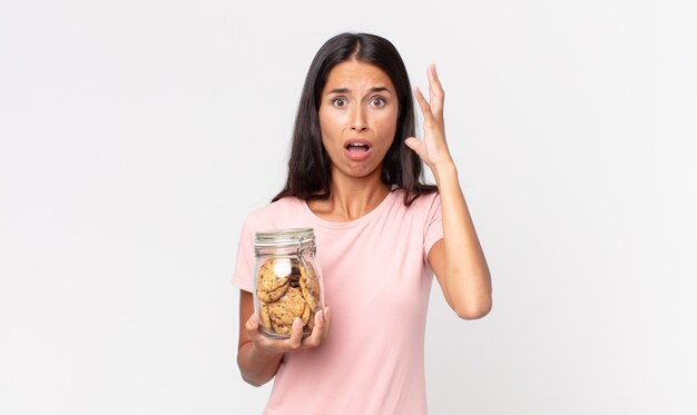 Young hispanic woman screaming with hands up in the air and holding a cookies glass bottle