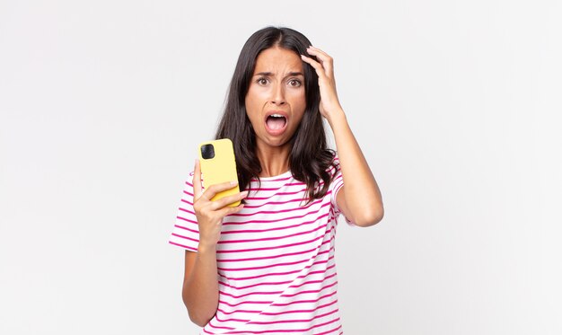 Young hispanic woman screaming with hands up in the air and holding a smartphone