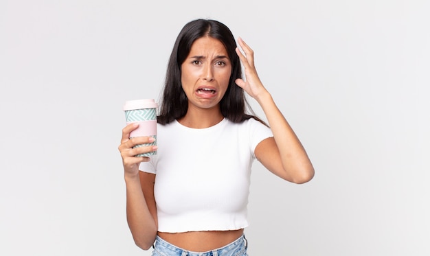 Young hispanic woman screaming with hands up in the air and holding a take away coffee container